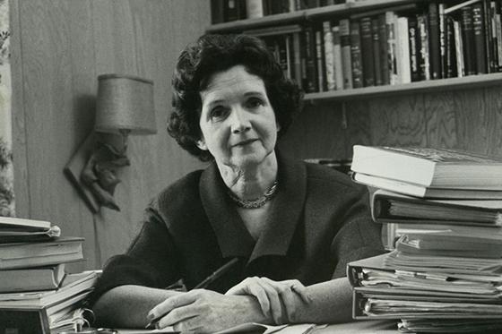 雷切尔·卡森的黑白照片，她坐在一张满是书的桌子后面，手里拿着笔. 